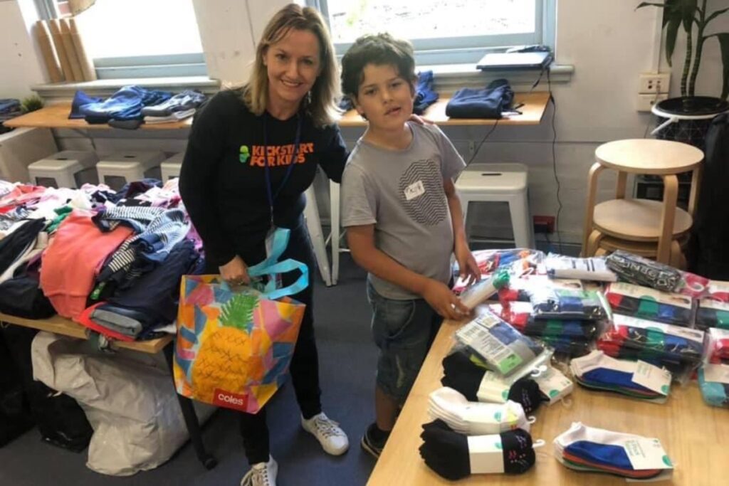 A woman in a KickStart for Kids t-shirt stands with a boy next to tables of clothing and packaged socks