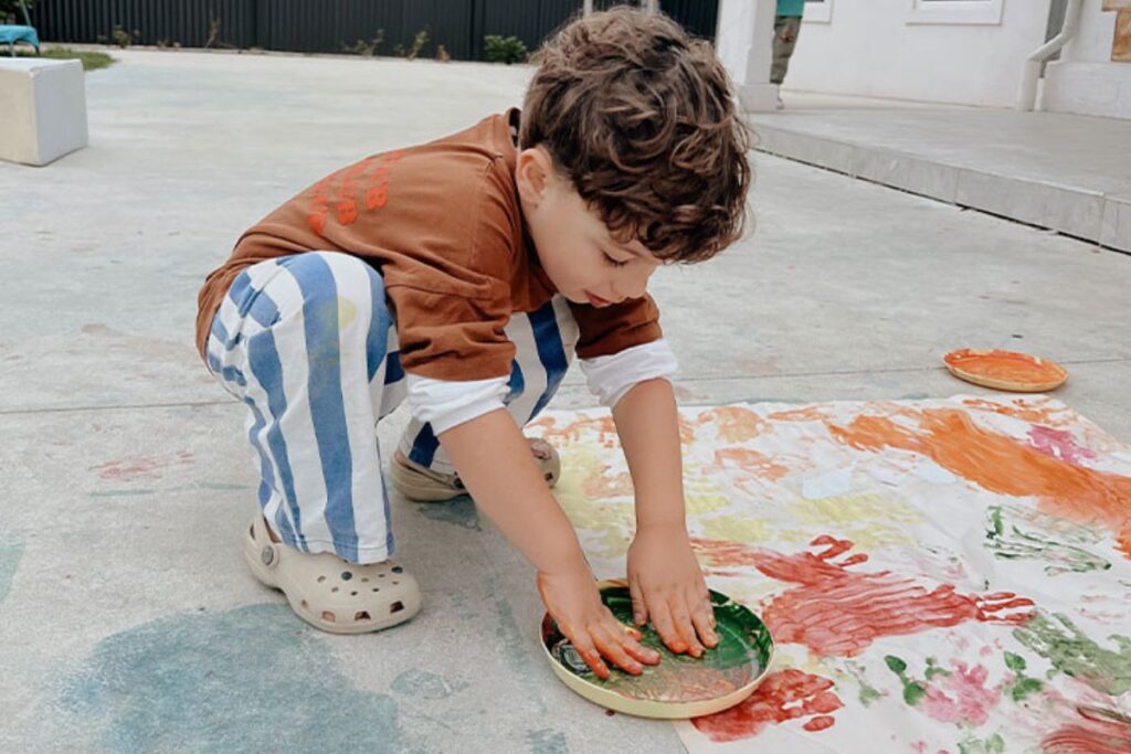 A young boy in a striped shirt and orange top kneels to finger paint on a large sheet spread across a concrete floor, with other paints nearby.