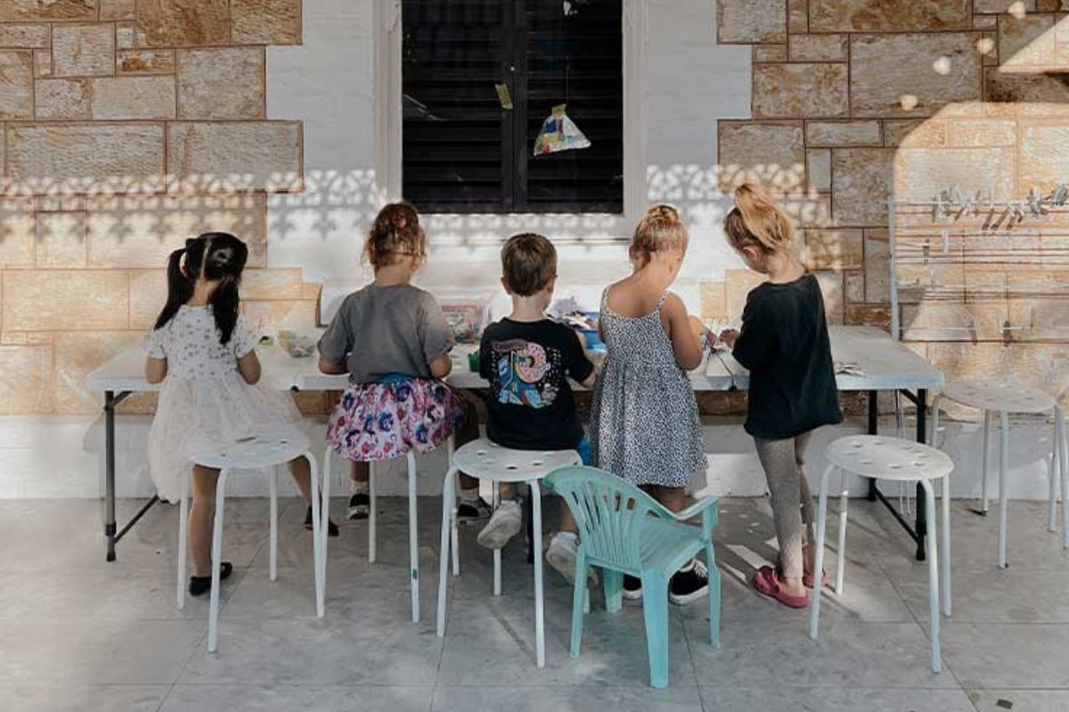 A group of young children stand at low tables in a studio, working together on a creative activity.