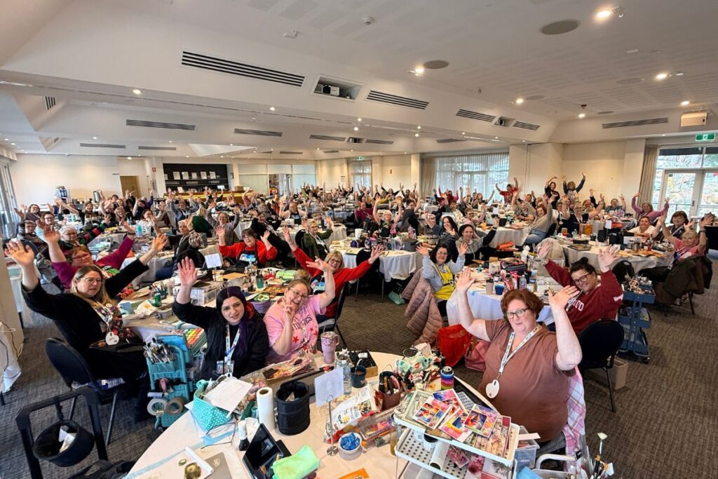 Large group of women with hands raised at a Natalie May scrapbooking event.