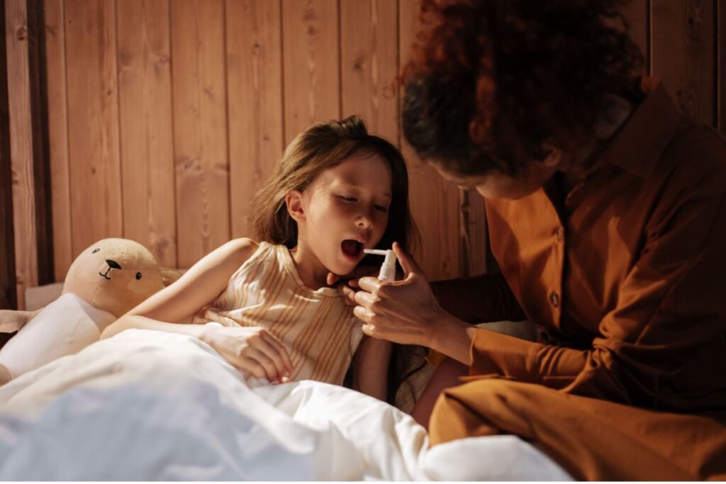 Parent administering throat spray to a child in bed with a teddybear.