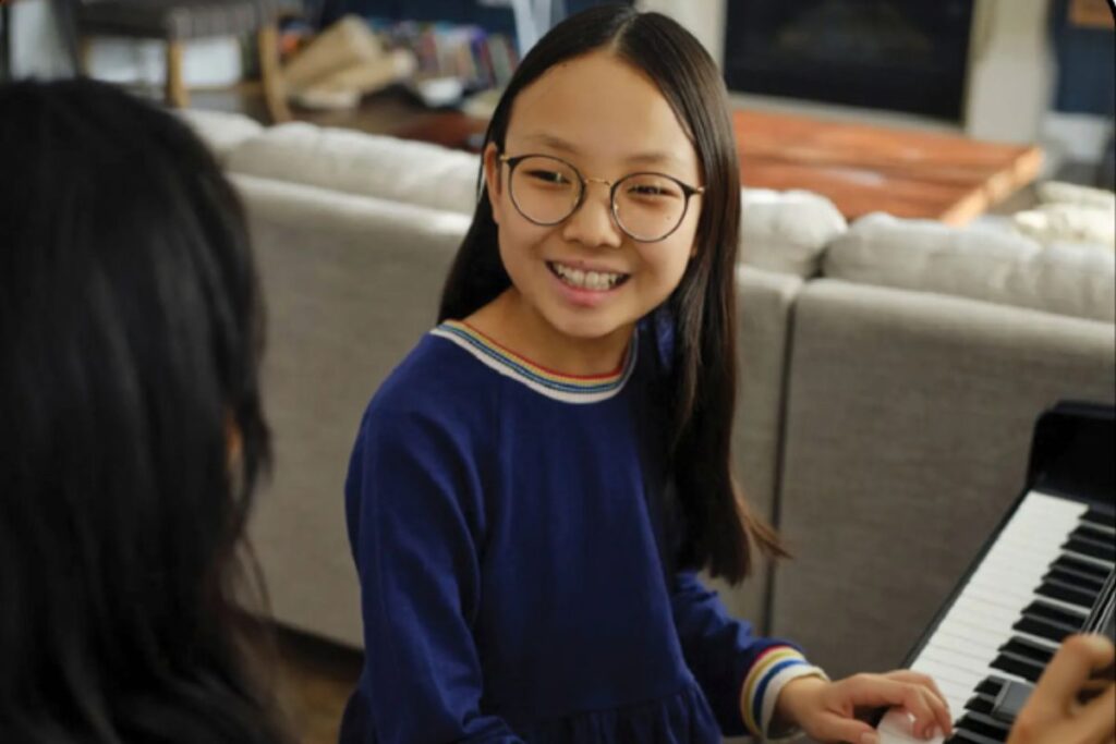 Girl in glasses smiling while playing piano during a music lesson, illustrating the cost of after-school extracurricular activities
