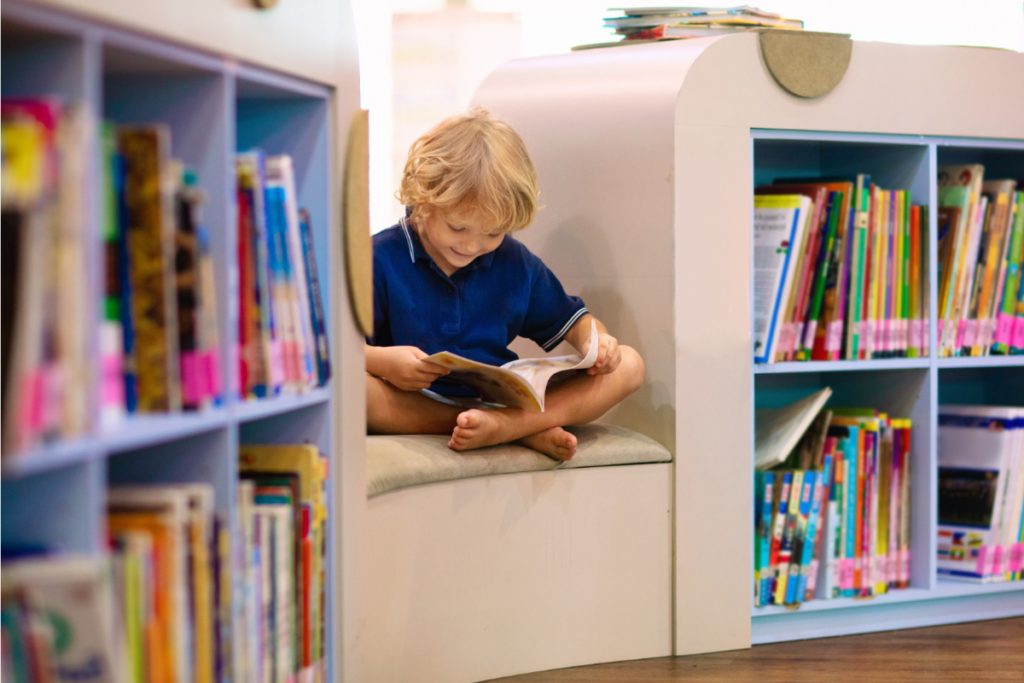 Young boy in school uniform sitting cross-legged in a library reading nook, reading a book surrounded by colourful bookshelves.